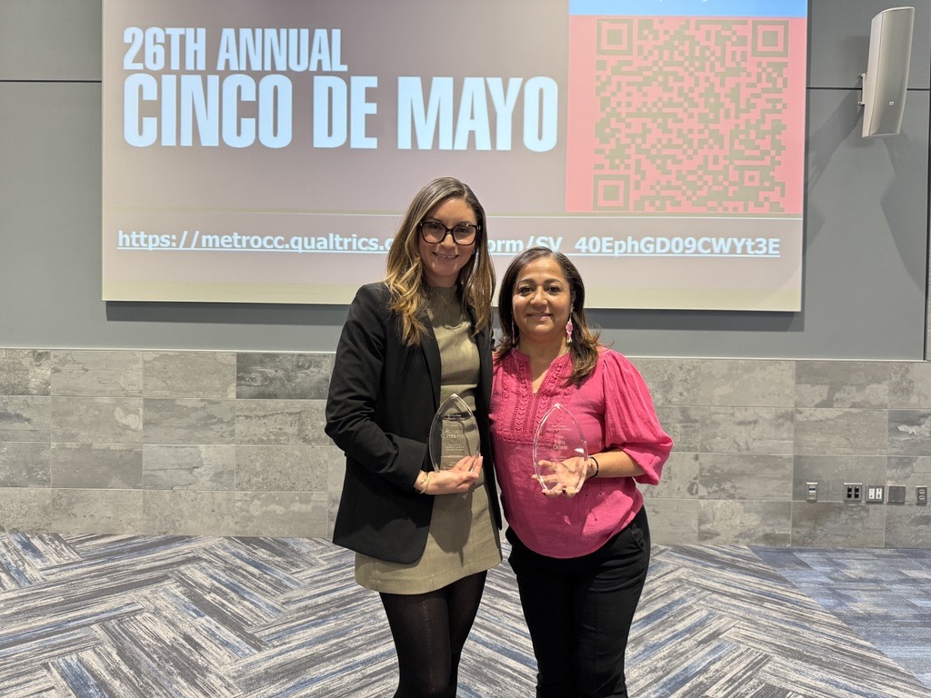 Two women stand smiling together at the 26th Annual Cinco de Mayo Celebration while holding crystal awards from Metropolitan Community College. A presentation screen with “Cinco de Mayo” text is visible in the background.