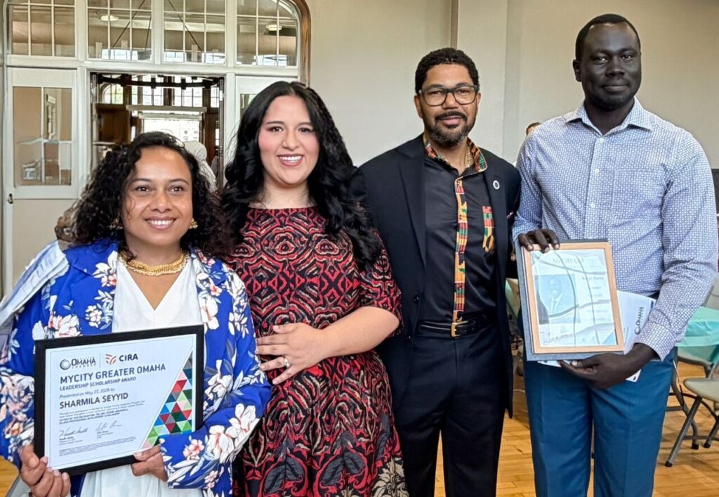 Four people stand together at a graduation celebration for the MyCity Greater Omaha leadership program. Two participants hold framed certificates while smiling in a bright indoor venue with large windows and wooden floors.