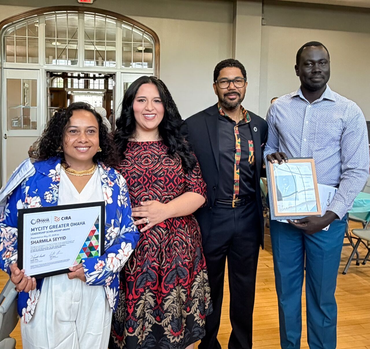 Four people stand together at a graduation celebration for the MyCity Greater Omaha leadership program. Two participants hold framed certificates while smiling in a bright indoor venue with large windows and wooden floors.